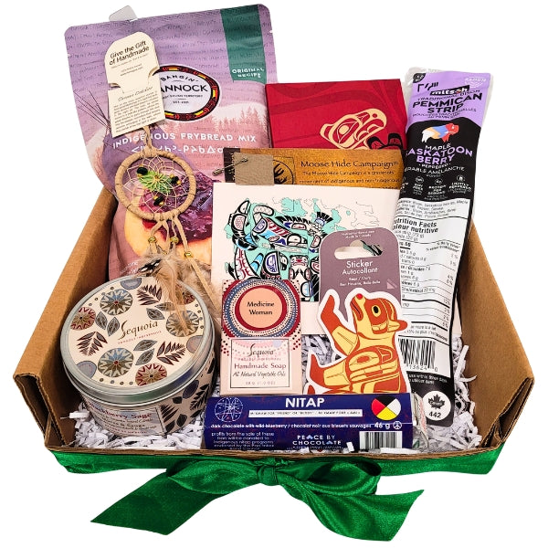 A gift box open to display various items including a dream catcher, a blackberry and sage soy candle, a eucalyptus soap, a moose hide pin, a Thunderbird and Orca notebook, a Nitap chocolate bar, and other food items.