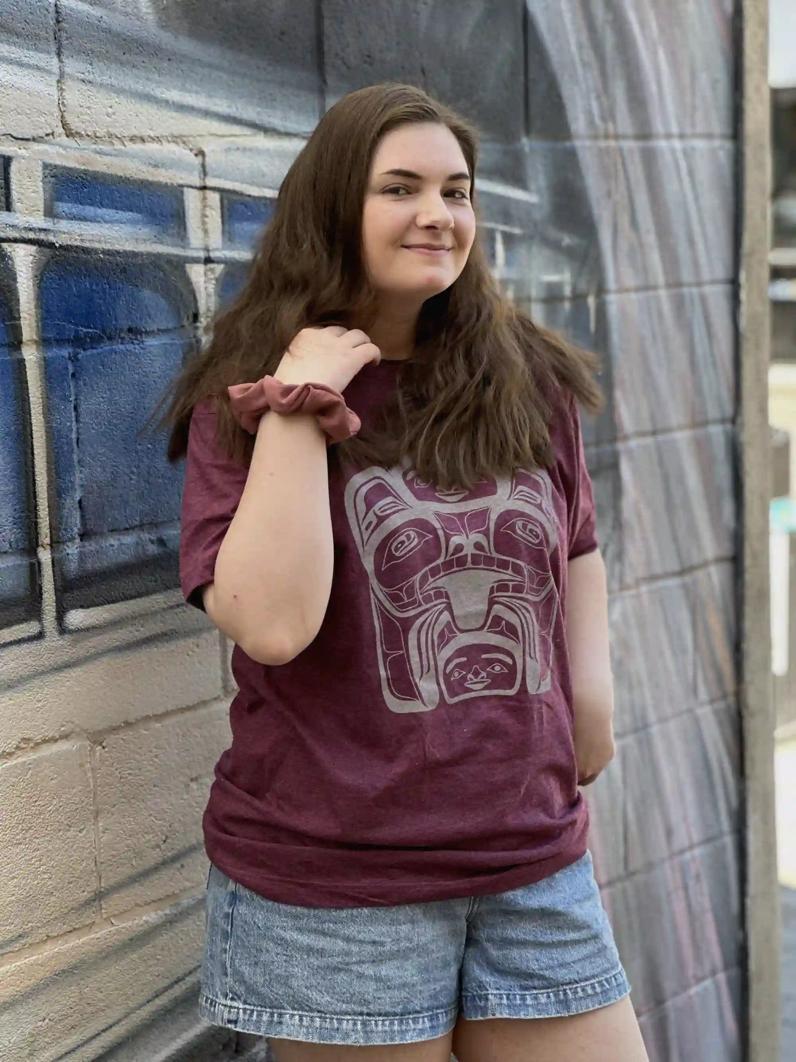 A woman with long brown hair stands against a brick wall with a painted mural, wearing a bear mother t-shirt.