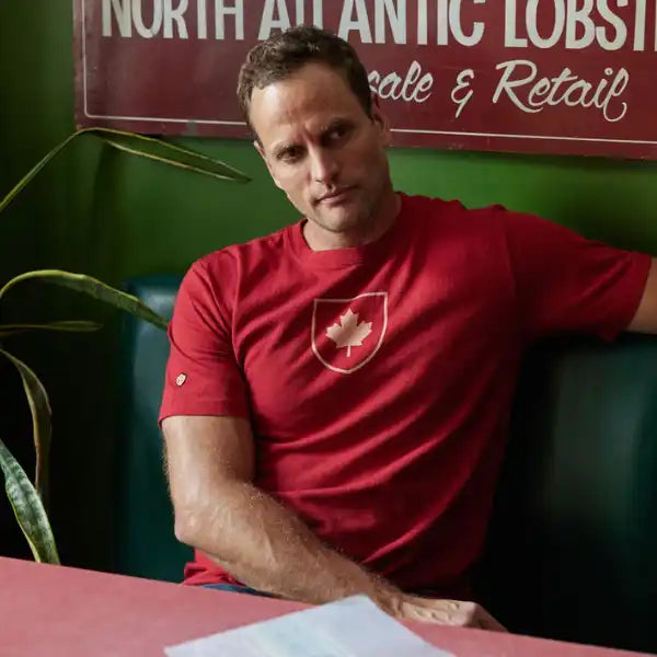A man wearing a red crewneck t-shirt with a white shield logo featuring a maple leaf, sitting in a green booth in front of a restaurant sign.