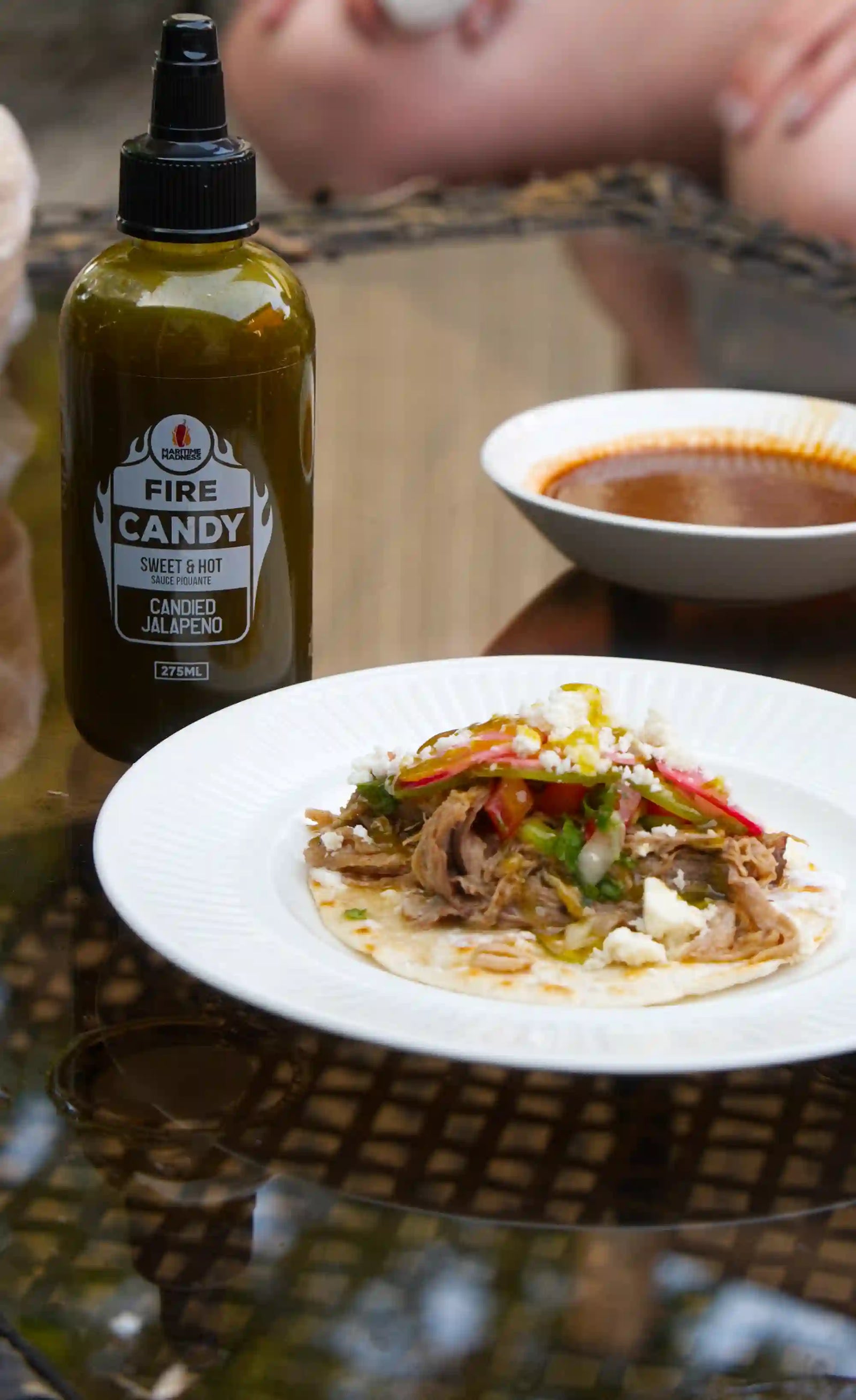 A bottle of Fire Candy Candied Jalapeño sauce next to a plate of prepared food on a table, with a blurred bowl in the background.