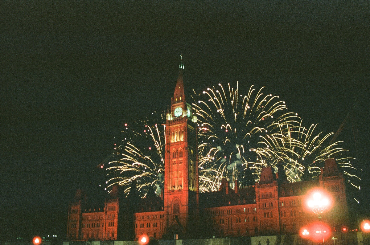 Fireworks exploding over the Centre Block of Canada's Parliament buildings. The front of the building is lit up with red light. 