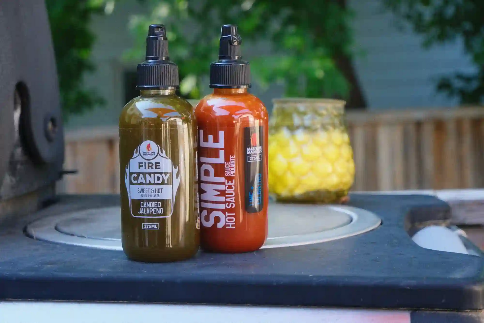 Two squeeze bottles of sauce standing on a dark tabletop outdoors, with a blurred background showing greenery and a fence. The left bottle is amber-brown with a black cap and a white label reading FIRE CANDY SPICED SAUCE.