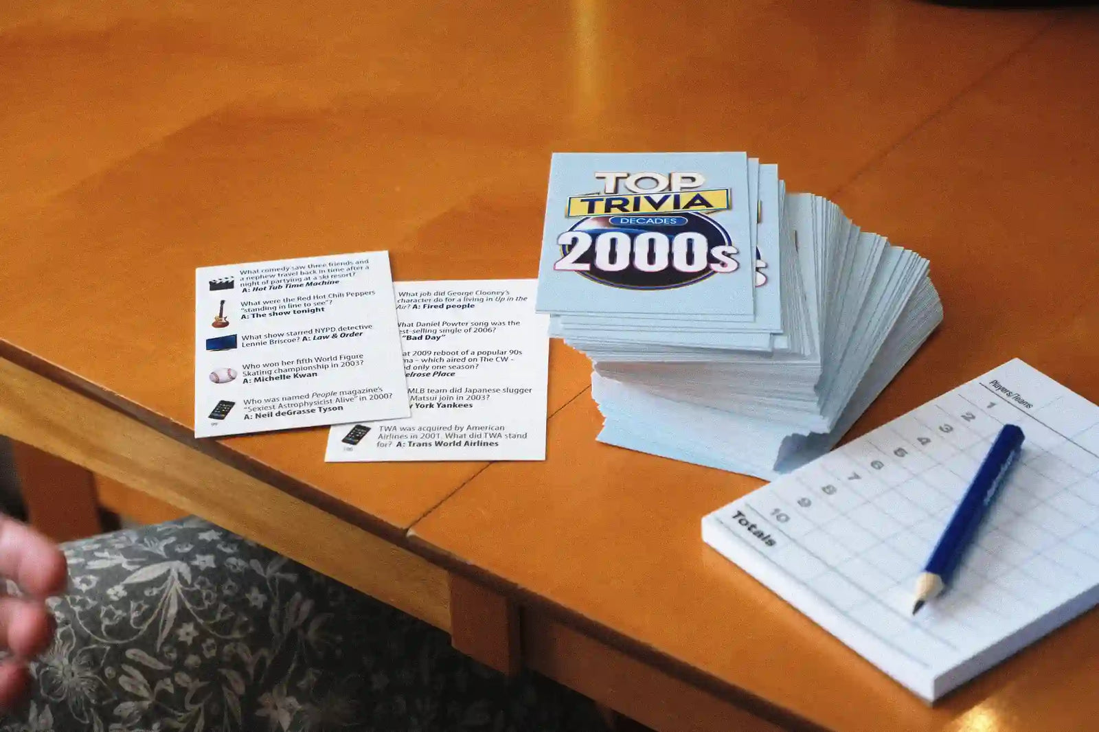 A stack of trivia cards titled "TOP TRIVA for the 2000s" sits on a wooden table next to a score sheet and pencil, with a hand partially visible at the lower left.