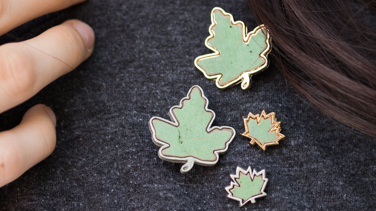 Four Under One Roof pins and brooches adorn a dark shirt, a woman's hair and hands enter at the corner of the frame. The pins and brooches feature the green patina copper of Canada's Parliament buildings. 