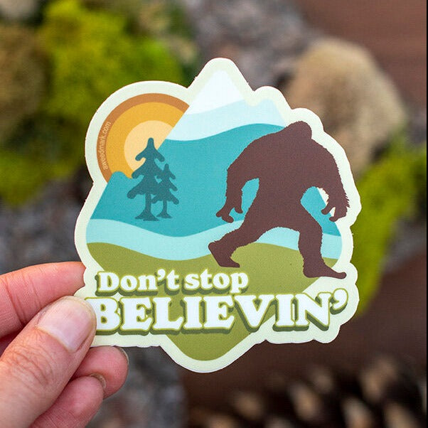 A hand holding a don't stop believin' big foot sticker.