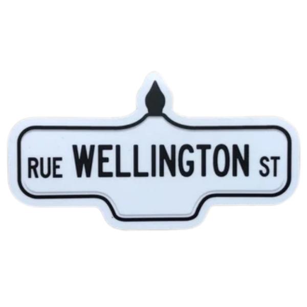 Sticker of a white street sign with black text that reads 'Rue Wellington St'.