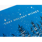 Card with blue background showing an illustration of a cozy winter A-frame cabin nestled in the trees. Snow is on the ground and falling from the sky. Silver text reads "Cozy Holiday Wishes".