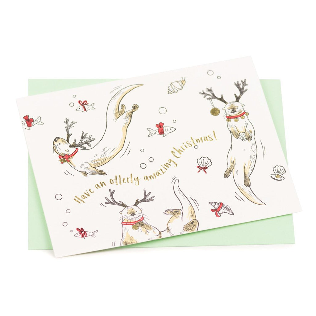A white card with illustrated playful otters wearing reindeer antlers and red collars. Gold foil text reads "Have an otterly amazing Christmas!"