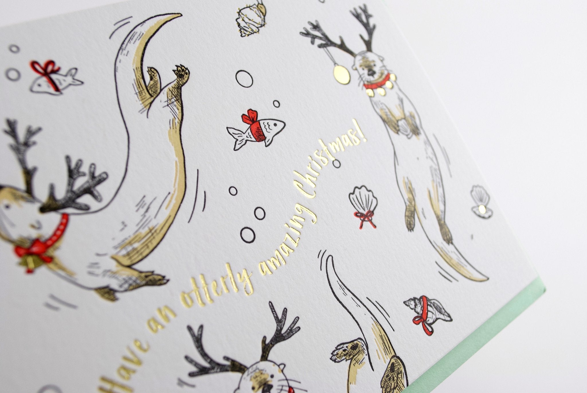 A white card with illustrated playful otters wearing reindeer antlers and red collars. Gold foil text reads "Have an otterly amazing Christmas!"