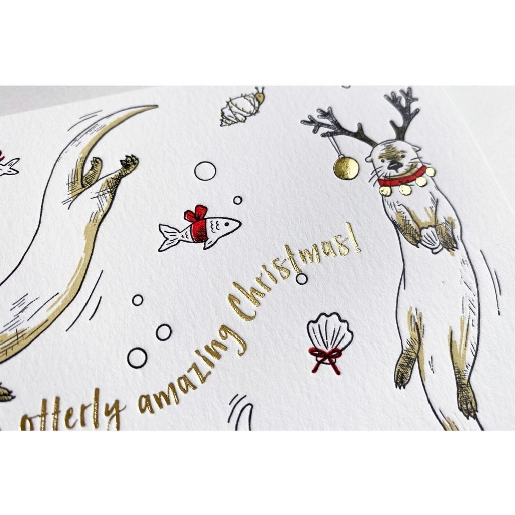 A white card with illustrated playful otters wearing reindeer antlers and red collars. Gold foil text reads "Have an otterly amazing Christmas!"