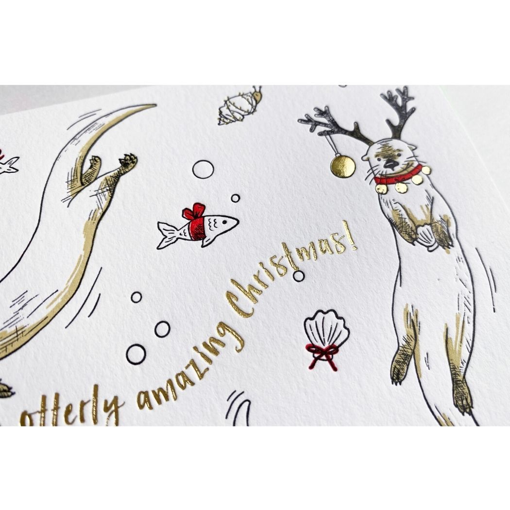 A white card with illustrated playful otters wearing reindeer antlers and red collars. Gold foil text reads "Have an otterly amazing Christmas!"